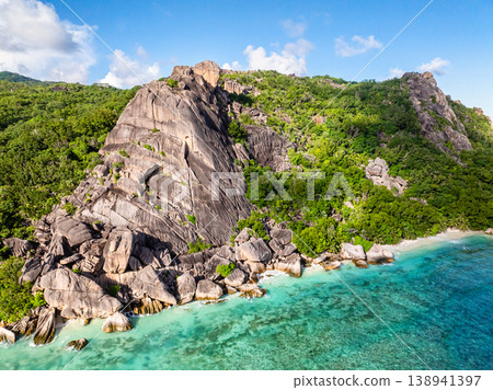 Imposing rocky cliffs rise above lush greenery, with turquoise waters at the base. Seychelles, La Digue. Imposing rocky cliffs rise above lush greenery, with turquoise waters at the base. Seychelles, La Digue. 138941397