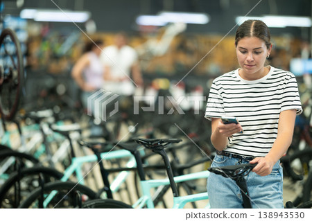 Young girl buying new bike in bicycle sports store, using smartphone to scan barcode of products Young girl buying new bike in bicycle sports store, using smartphone to scan barcode of products 138943530