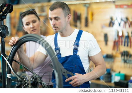 Positive man seller wearing uniform fixing bike and talking to female client in sport store Positive man seller wearing uniform fixing bike and talking to female client in sport store 138943531