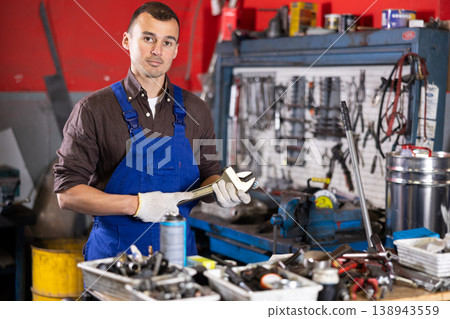 Positive male worker of car workshop holding metal tool in car workshop Positive male worker of car workshop holding metal tool in car workshop 138943559