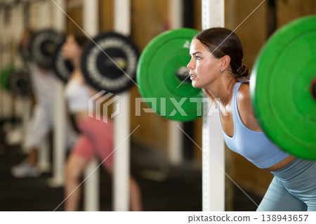 Motivated crossfit female athlete lifting heavy body bar or barbell during group training in sport gym Motivated crossfit female athlete lifting heavy body bar or barbell during group training in sport gym 138943657
