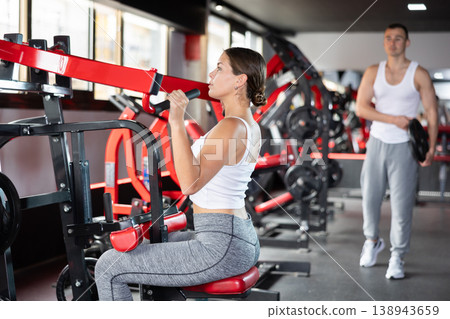 Girl doing strength training in gym, exercising upper body muscles on shoulder press machine Girl doing strength training in gym, exercising upper body muscles on shoulder press machine 138943659