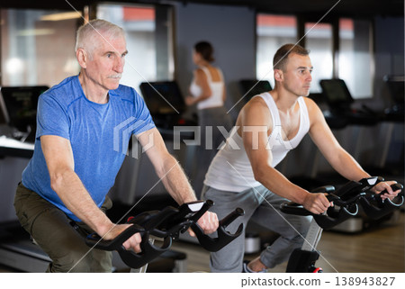 Two men exercising on stationary exercise bikes in gym 138943827