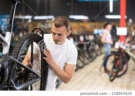 Young male customer choosing bike and checking tire on bicycle wheel in sport store 138943828