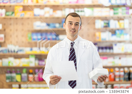 Young male pharmacist standing in drugstore with medicament and recipe in hands Young male pharmacist standing in drugstore with medicament and recipe in hands 138943874