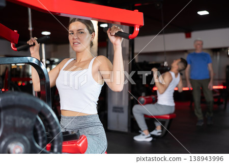 Girl doing strength training in gym, exercising upper body muscles on shoulder press machine Girl doing strength training in gym, exercising upper body muscles on shoulder press machine 138943996