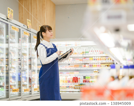 A female store clerk checking a tablet in the aisle of a supermarket. 138945009