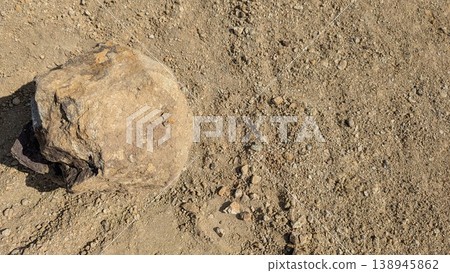 Large natural rock sits on dry, sandy soil. Grainy earth texture surrounds the solid stone. High angle view highlights the contrast between the heavy mineral, parched ground. Minimalist landscape. 138945862