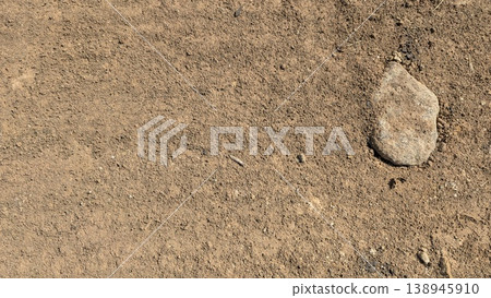 Natural dry soil surface displays a rough, grainy texture. A solitary rock rests on the parched ground. Earth tones dominate this minimalist landscape. High angle view shows raw, organic details. Natural dry soil surface displays a rough, grainy texture. A solitary rock rests on the parched ground. Earth tones dominate this minimalist landscape. High angle view shows raw, organic details. 138945910