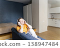 Smiling woman enjoying audio with wireless headphones, sitting comfortably on a sofa in a modern apartment. Woman relaxing at home, listening to music 138947484