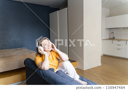 Smiling woman enjoying audio with wireless headphones, sitting comfortably on a sofa in a modern apartment. Woman relaxing at home, listening to music 138947484