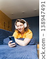 Woman wearing headphones and using a smartphone while relaxing on a blue sofa, enjoying leisure time and entertainment, surfing the internet on his phone, listening to music with headphones 138947491