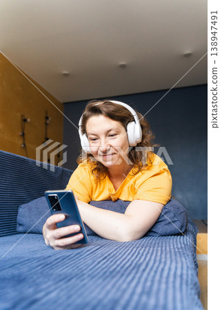 Woman wearing headphones and using a smartphone while relaxing on a blue sofa, enjoying leisure time and entertainment, surfing the internet on his phone, listening to music with headphones Woman wearing headphones and using a smartphone while relaxing on a blue sofa, enjoying leisure time and entertainment, surfing the internet on his phone, listening to music with headphones 138947491