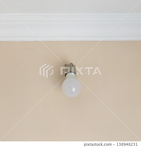 Ceiling light bulb mounted on plain wall in indoor space during daytime with natural light coming through 138948231