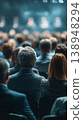 Many people sitting in a large hall attending a conference during daytime with speakers visible in the background 138948294