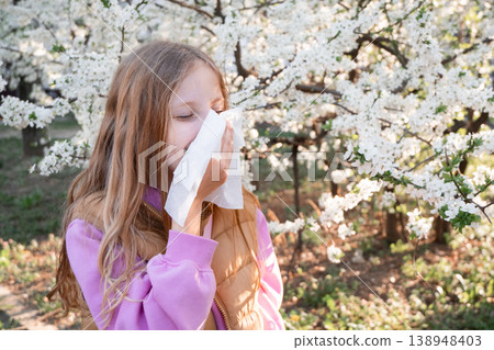 Young girl blowing her nose into a tissue near blooming trees. Child spring allergy, hay fever symptom, outdoor health issue. Young girl blowing her nose into a tissue near blooming trees. Child spring allergy, hay fever symptom, outdoor health issue. 138948403
