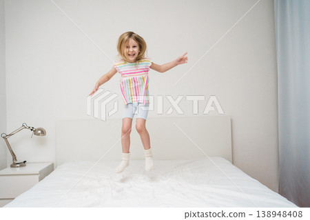 A smiling girl jumps on a bed with arms spread out in a bright minimalist room wearing a colorful rainbow striped shirt joyful energetic childhood play home family lifestyle concept 138948408