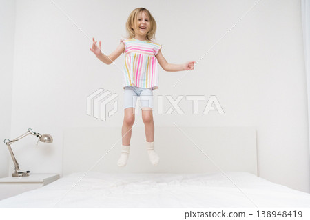 A smiling girl jumps on a bed with arms spread out in a bright minimalist room wearing a colorful rainbow striped shirt joyful energetic childhood play home family lifestyle concept 138948419