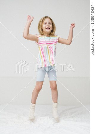 A smiling girl jumps on a bed with arms spread out in a bright minimalist room wearing a colorful rainbow striped shirt joyful energetic childhood play home family lifestyle concept A smiling girl jumps on a bed with arms spread out in a bright minimalist room wearing a colorful rainbow striped shirt joyful energetic childhood play home family lifestyle concept 138948424
