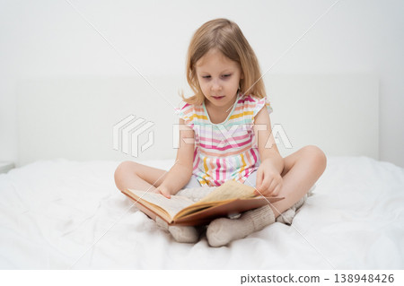A smiling girl sits cross-legged on a bed reading a large book with both hands in a bright minimalist room cozy home learning childhood literacy education family lifestyle concept A smiling girl sits cross-legged on a bed reading a large book with both hands in a bright minimalist room cozy home learning childhood literacy education family lifestyle concept 138948426