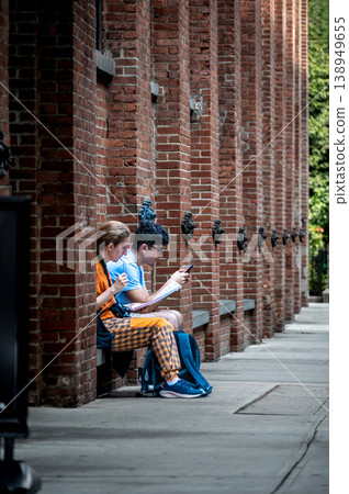 New york, usa august 15,2024. Two young friends sit on a brooklyn street bench by a brick wall, using smartphones and a map while exploring dumbo, nyc 138949655