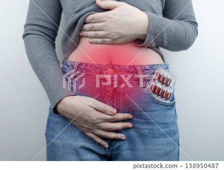 Close-up of a woman suffering from menstrual cramps holding her belly, with painkillers in her jeans pocket. Female health, period pain relief, and abdominal ache concept. Close-up of a woman suffering from menstrual cramps holding her belly, with painkillers in her jeans pocket. Female health, period pain relief, and abdominal ache concept. 138950487
