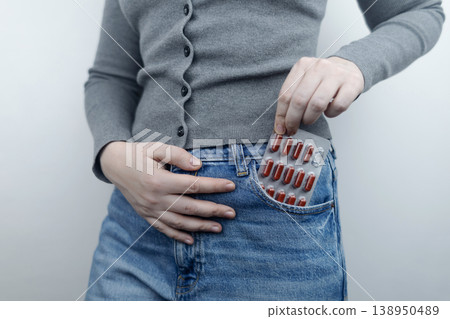 Close-up of a woman suffering from menstrual cramps holding her belly, with painkillers in her jeans pocket. Female health, period pain relief, and abdominal ache concept. Close-up of a woman suffering from menstrual cramps holding her belly, with painkillers in her jeans pocket. Female health, period pain relief, and abdominal ache concept. 138950489