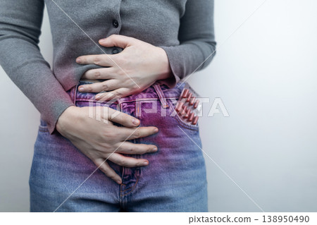 Close-up of a woman suffering from menstrual cramps holding her belly, with painkillers in her jeans pocket. Female health, period pain relief, and abdominal ache concept. Close-up of a woman suffering from menstrual cramps holding her belly, with painkillers in her jeans pocket. Female health, period pain relief, and abdominal ache concept. 138950490