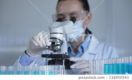 A scientist woman, wearing a lab coat, mask, white gloves and safety glasses, is adjusting a microscope surrounded by test tubes in laboratory A scientist woman, wearing a lab coat, mask, white gloves and safety glasses, is adjusting a microscope surrounded by test tubes in laboratory 138950543