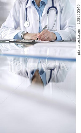 Unknown doctor woman, wearing a stethoscope and blue blouse under a lab coat, is using a clipboard and taking notes on glass desk in a medical office, close-up vertical view. Medicine and science Unknown doctor woman, wearing a stethoscope and blue blouse under a lab coat, is using a clipboard and taking notes on glass desk in a medical office, close-up vertical view. Medicine and science 138950546