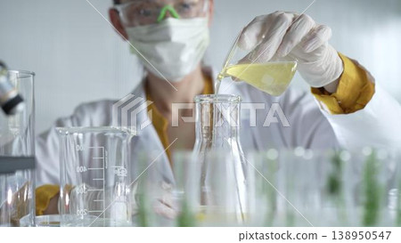 Female scientist researcher wearing a lab coat, white gloves, and mask, is pouring a yellow oily liquid from a beaker into an Erlenmeyer flask in laboratory, portrait. Science and medicine concepts 138950547