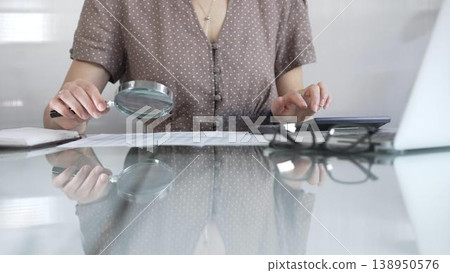 Businesswoman accountant in brawn dotted blouse reviewing financial reports with magnifying glass and calculator, conducting audit and analyzing data in office. Taxes, audit in business 138950576