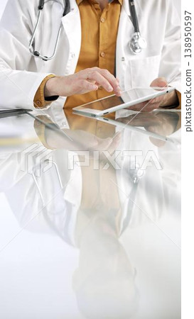 Unknown male doctor wearing a stethoscope, yellow shirt under a lab coat, is using digital tablet in modern healthcare setting, interacting with patient data, vertical portrait. Medicine concept 138950597