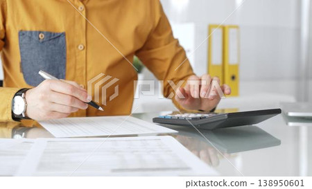 Professional calculating finances using a desktop calculator and writing on tax documents in an office setting. Audit and taxes in business Professional calculating finances using a desktop calculator and writing on tax documents in an office setting. Audit and taxes in business 138950601