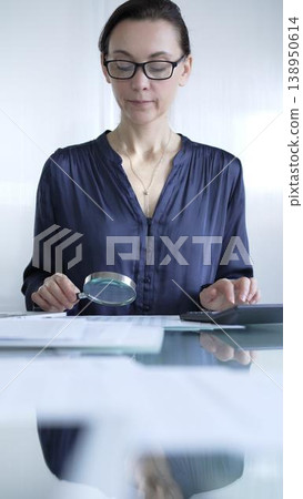 Woman accountant examining financial documents with magnifying glass at glass office desk, conducting thorough analysis of business transactions. Audit and taxes in business 138950614