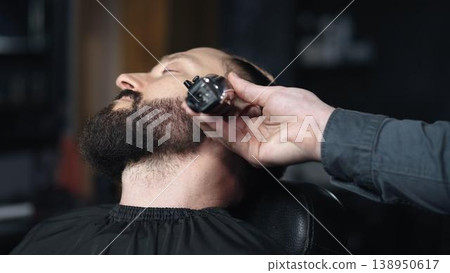 Professional barber trimming client's beard with electric razor, delivering precise styling within contemporary grooming salon setting Professional barber trimming client's beard with electric razor, delivering precise styling within contemporary grooming salon setting 138950617