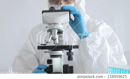 A male doctor in a protective suit and gloves uses a microscope while analyzing samples, conducting research in a laboratory. Close-up of test tubes with blue liquid A male doctor in a protective suit and gloves uses a microscope while analyzing samples, conducting research in a laboratory. Close-up of test tubes with blue liquid 138950625