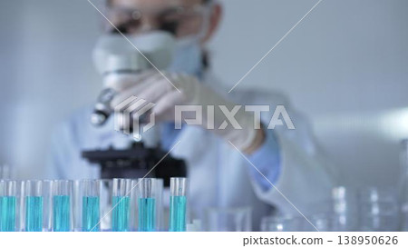 Laboratory tubes, filled with a blue liquid, are in focus, closeup view. Female scientist researcher wearing white gloves and protective mask is using a microscope at the background Laboratory tubes, filled with a blue liquid, are in focus, closeup view. Female scientist researcher wearing white gloves and protective mask is using a microscope at the background 138950626