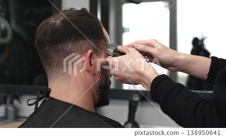 Professional barber trimming male client's hair with electric razor, delivering precise grooming service in contemporary, well lit barbershop setting 138950641