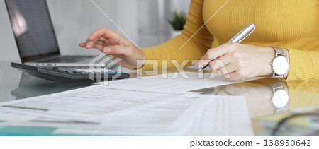 Businesswoman wearing yellow casual sweater is calculating budget and reviewing financial documents using a laptop and calculator for home finances. Audit and taxes in business Businesswoman wearing yellow casual sweater is calculating budget and reviewing financial documents using a laptop and calculator for home finances. Audit and taxes in business 138950642