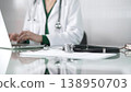 Female doctor using technology in healthcare, close up of hands typing on laptop with stethoscope, glasses, and clipboard on desk 138950703