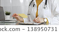Doctor wearing stethoscope and yellow sweater under white lab coat, making notes on a clipboard in a bright medical office. Medicine, healthcare and science concept 138950731