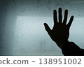 Dark silhouette of a hand pressed against a wet, condensed glass surface in cool blue tones. Conceptual image symbolizing loneliness, separation, or a plea for help through a foggy window with water 138951002