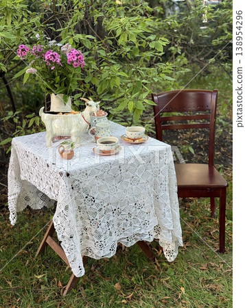 Tea set on ornate tablecloth in garden setting, featuring flowers and wooden chair background Tea set on ornate tablecloth in garden setting, featuring flowers and wooden chair background 138954296