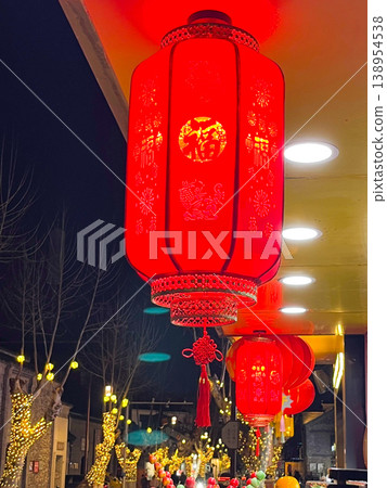 Glowing cylindrical red fabric lanterns with tassels hanging outdoors at night near street trees covered in lights, for Lunar New Year and Asian festival themes. 138954538