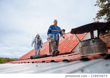 Two men repairs chimney on house roof. Roofer working on metal rooftop construction for home maintenance. Professional worker repairing ventilation system. Building improvement service. Two men repairs chimney on house roof. Roofer working on metal rooftop construction for home maintenance. Professional worker repairing ventilation system. Building improvement service. 138954674