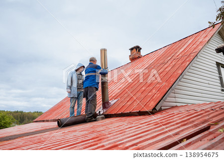 Man and woman repairing chimney on red metal roof of house. Experienced technician installing new steel flue pipe system. Maintenance work at height for heating safety and ventilation. Man and woman repairing chimney on red metal roof of house. Experienced technician installing new steel flue pipe system. Maintenance work at height for heating safety and ventilation. 138954675