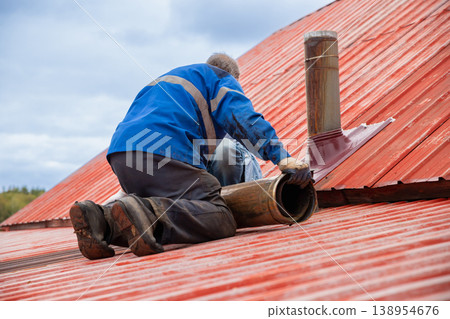 Chimney sweep worker installing pipe on red metal roof. Roofer fixing flue system on house exterior. Repair of heating ventilation and construction maintenance concept. Professional builder. Chimney sweep worker installing pipe on red metal roof. Roofer fixing flue system on house exterior. Repair of heating ventilation and construction maintenance concept. Professional builder. 138954676