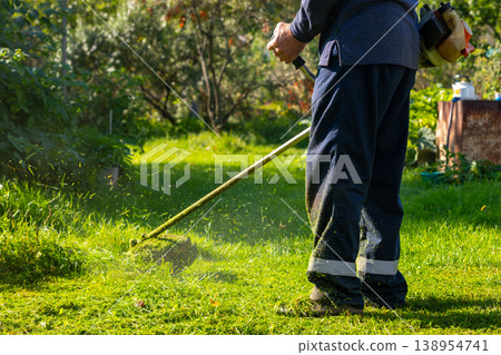 Man using grass trimmer to cut lawn in backyard. Gardener operating string trimmer to maintain greenery. Lawn care equipment, landscaping tool and grass maintenance gardening concept. Man using grass trimmer to cut lawn in backyard. Gardener operating string trimmer to maintain greenery. Lawn care equipment, landscaping tool and grass maintenance gardening concept. 138954741