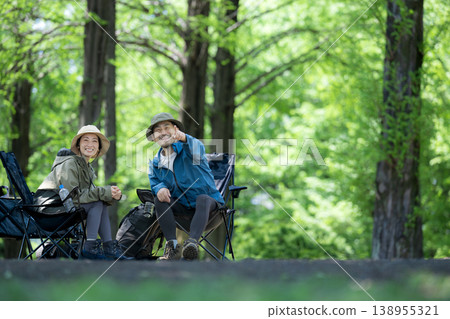 一對面帶微笑的中年夫婦在綠樹環繞的露營地裡，坐在椅子上享受戶外休閒活動，觀賞鳥類和風景。 138955321